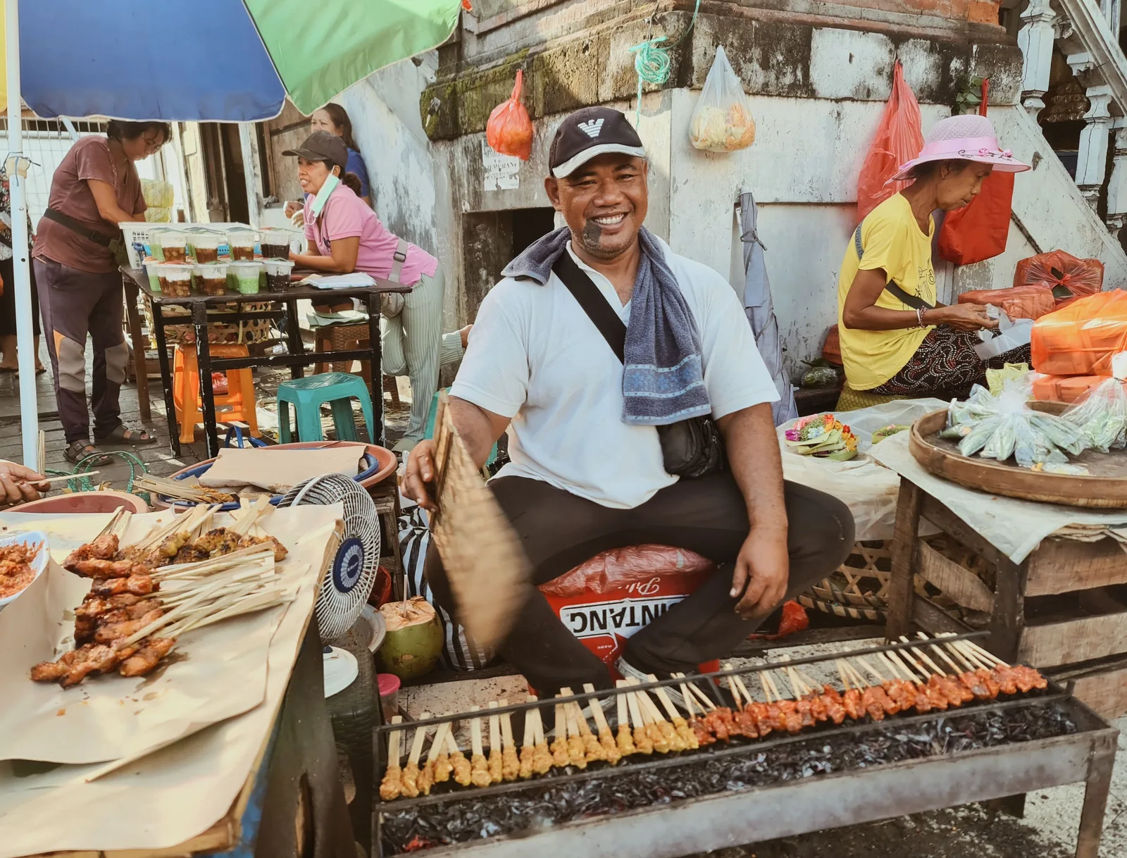 Satay vendor grilling skewers at a market in Bali, with a crate of Bintang beer visible